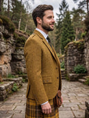 A man in profile wearing a mustard-yellow tweed blazer, a yellow-and-brown plaid kilt, a white shirt, and a dark tie, standing outdoors on a stone path with trees and rock walls in the background.