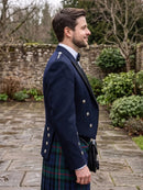 A man in profile wearing a navy blue formal jacket with silver buttons and a green tartan kilt, standing outdoors on a stone path with shrubs and a stone wall in the background.