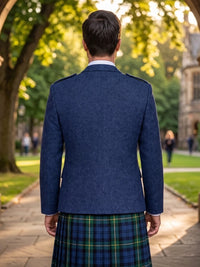 A man viewed from behind wearing a navy blue tweed blazer and a green and blue tartan kilt, standing on a stone path in a sunny park with trees and a historic building in the background.