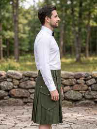 A man wearing a white long-sleeve dress shirt and a dark olive green pleated kilt, standing outdoors in front of a stone wall and a green forest background.