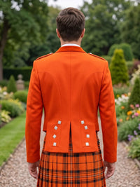 A man viewed from behind wearing a bright orange Scottish kilt jacket with silver buttons and a matching orange and black plaid kilt, standing in a garden setting.