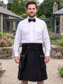 A man wearing a black kilt with silver hardware, a white dress shirt, and a black bow tie, standing outdoors in front of a stone building and garden.