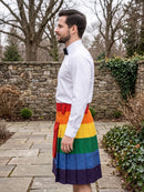A man in a white dress shirt and black bow tie stands in profile outdoors wearing a pleated kilt with horizontal rainbow stripes in red, orange, yellow, green, blue, and purple.