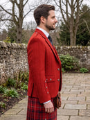 A man in profile wearing a bright red tweed blazer, a red and black tartan kilt, a white shirt, and a dark tie, standing outdoors on a stone path with a stone wall and bare trees in the background.
