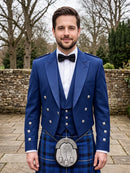 A man wearing a formal Scottish outfit, featuring a royal blue double-breasted jacket with silver buttons, a matching blue plaid kilt, a white shirt, a black bow tie, and a silver sporran, standing outdoors in front of a stone wall and trees.