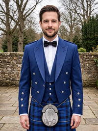 A man wearing a formal Scottish outfit, featuring a royal blue double-breasted jacket with silver buttons, a matching blue plaid kilt, a white shirt, a black bow tie, and a silver sporran, standing outdoors in front of a stone wall and trees.
