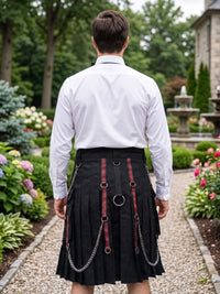 A man viewed from behind wearing a black pleated kilt with red and black patterned straps, silver metal rings, and hanging chains, paired with a white dress shirt in a garden setting.