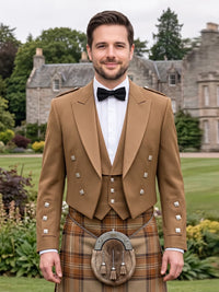 A man wearing a traditional Scottish formal outfit with a tan jacket, white shirt, black bow tie, and a brown tartan kilt with a sporran, standing outdoors in front of a stone mansion.