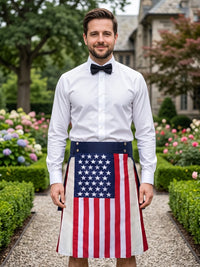 A man standing in a garden wearing a white dress shirt, black bow tie, and a kilt featuring the American flag pattern.