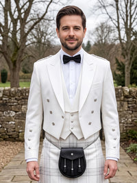 A man wearing a formal white double-breasted jacket with silver buttons, a white waistcoat, a white shirt, a black bow tie, and a black sporran on a white plaid kilt, standing outdoors in front of a stone wall and bare trees.