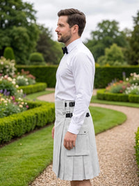 A man standing in profile in a formal garden, wearing a white long-sleeve dress shirt, black bow tie, and a light gray pleated kilt with black belt details.