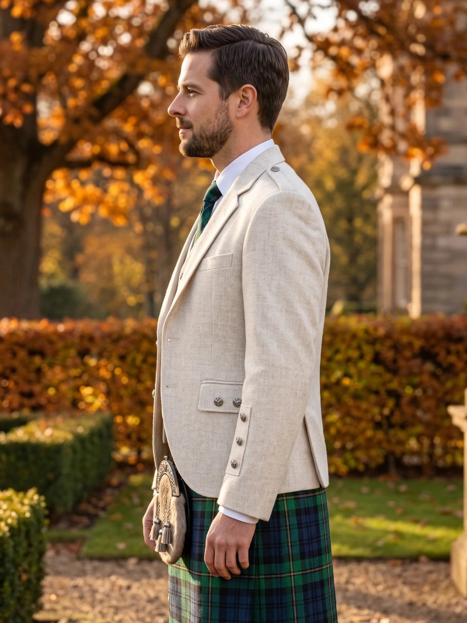 A man in profile wearing a light beige tweed blazer, a green and navy tartan kilt, a white dress shirt, and a green tie, standing outdoors in a garden with autumn foliage.