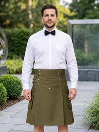 A man wearing a white dress shirt, black bow tie, and an olive green kilt with decorative metal rivets and flap pockets, standing outdoors on a paved walkway with greenery in the background.