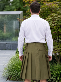 A man viewed from behind wearing a white long-sleeve button-up shirt and an olive green pleated kilt with side cargo pockets, standing outdoors in front of green plants.