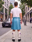 A man standing on a city sidewalk, viewed from behind, wearing a white t-shirt, a bright blue pleated kilt, white knee-high socks, and black shoes. Buildings, trees, a black Mercedes, and a bicycle are visible in the background.