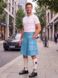 A man stands on a city sidewalk wearing a light blue kilt, white t-shirt, white knee-high socks, and black shoes. Buildings, trees, a black car, and a bicycle are visible in the background.