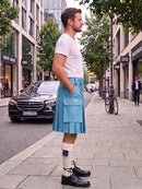 A man standing on a city sidewalk wearing a light blue pleated kilt, white t-shirt, white socks, and black leather shoes, with a street scene and parked cars in the background.