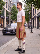 A man wearing a tan pleated kilt with red side panels, a white t-shirt, white knee-high socks, and black shoes, standing on a city sidewalk next to a street with a Mercedes-Benz and trees.