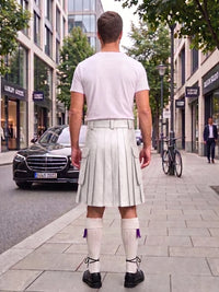 A man viewed from behind wearing a white short-sleeve t-shirt and a pleated white kilt, standing on a city sidewalk with buildings, cars, and a bicycle in the background.