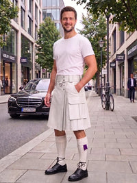 A man stands on a city sidewalk wearing a white short-sleeve t-shirt and a pleated white kilt-style skirt with a matching belt, paired with white knee-high socks and black lace-up shoes. Buildings, trees, a parked black car, and a bicycle are visible in the urban street setting.